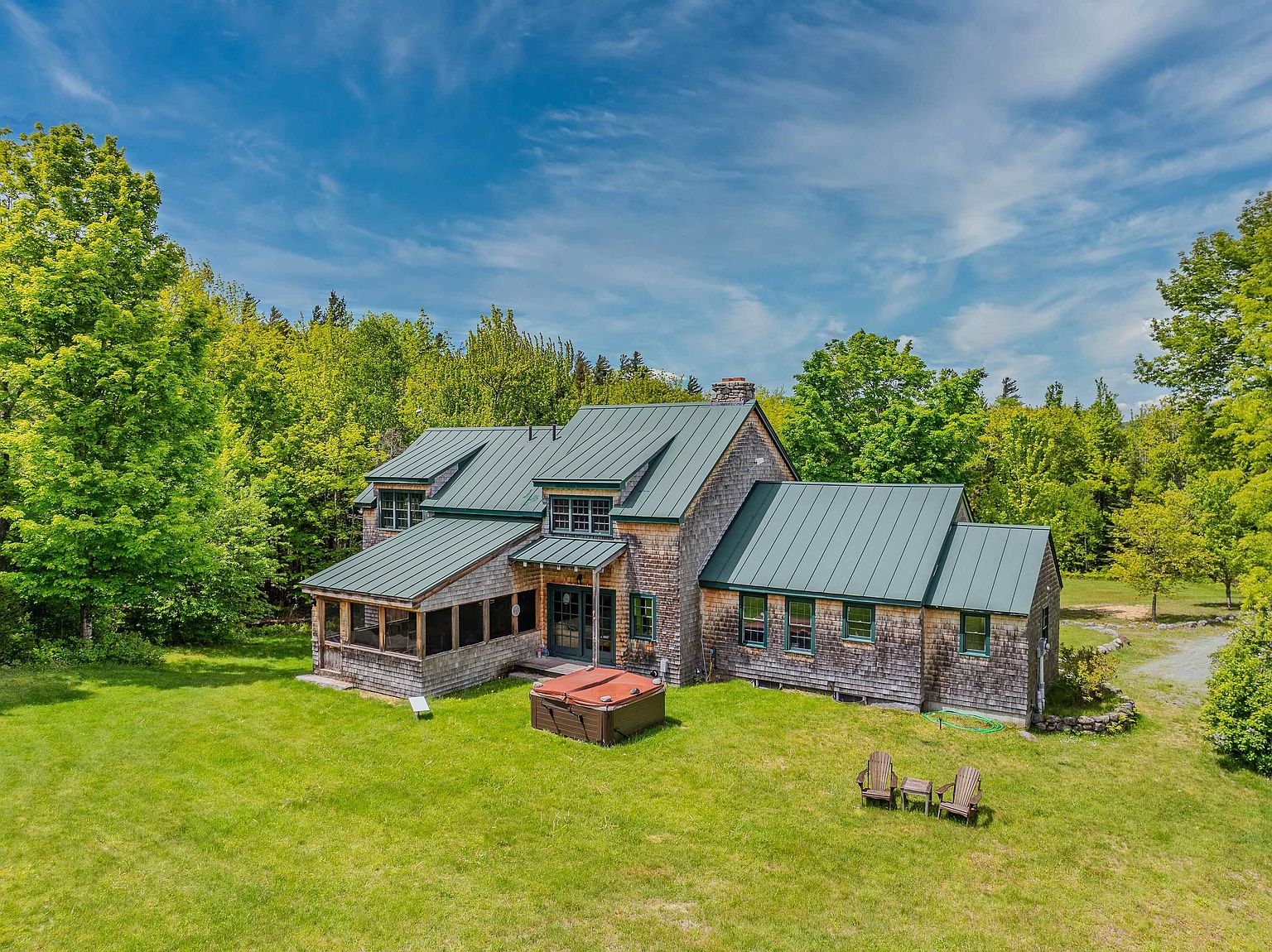 This aerial perspective captures a charming shingle-style home nestled against a lush, dense forest backdrop. The residence features a distinctive green metal roof, a screened-in porch, and a private hot tub situated on the expansive green lawn. Two wooden Adirondack chairs sit in the foreground, emphasizing the peaceful, secluded nature of this rural property.