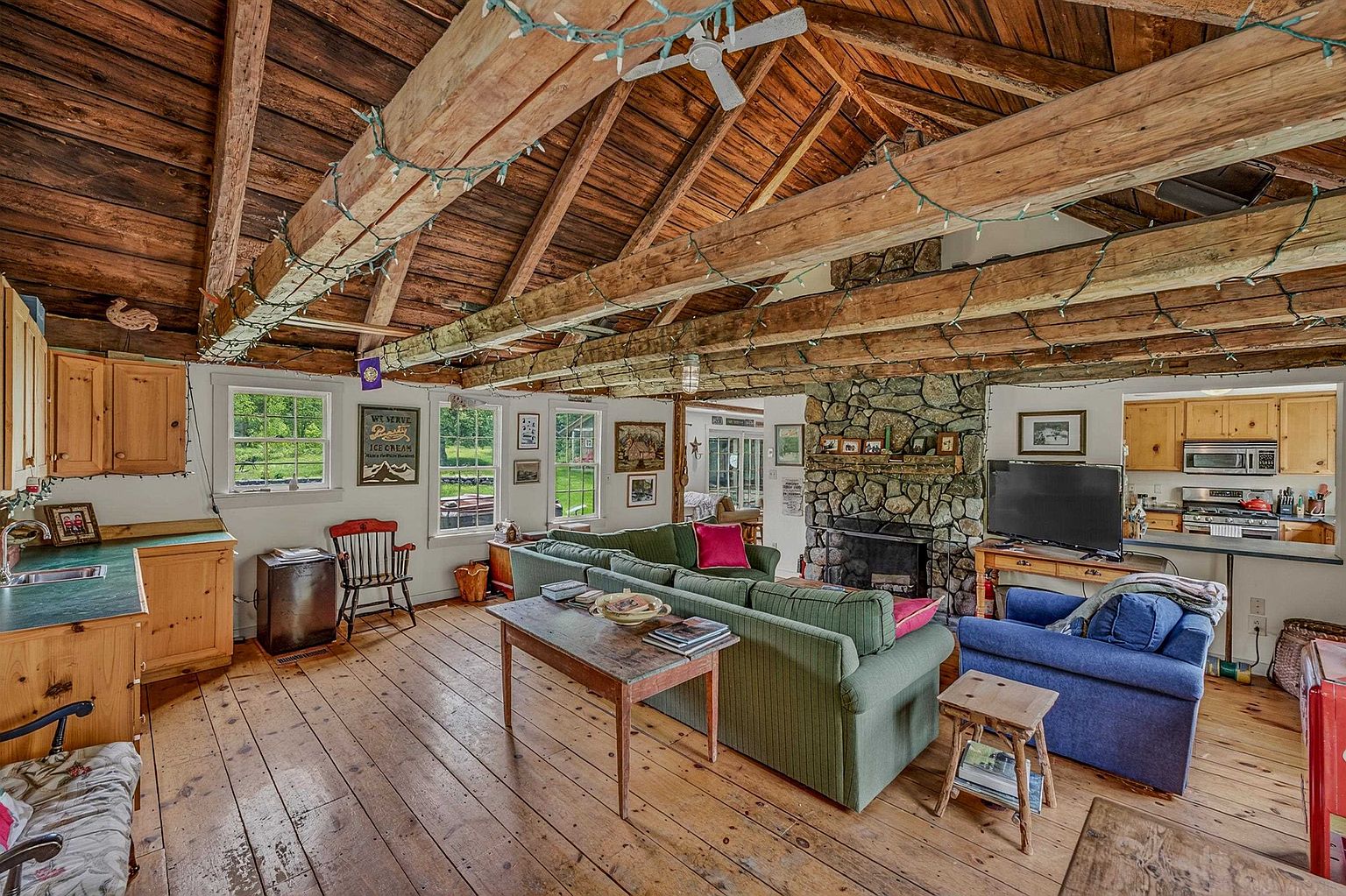 This rustic living room features exposed timber beams across a vaulted ceiling and warm wood flooring throughout. A prominent stone-clad chimney breast serves as a focal point, complemented by a green sectional sofa and a blue armchair arranged for comfortable seating. The open-concept layout flows into a kitchen area in the background, creating a cozy and inviting atmosphere with a cabin-like aesthetic.