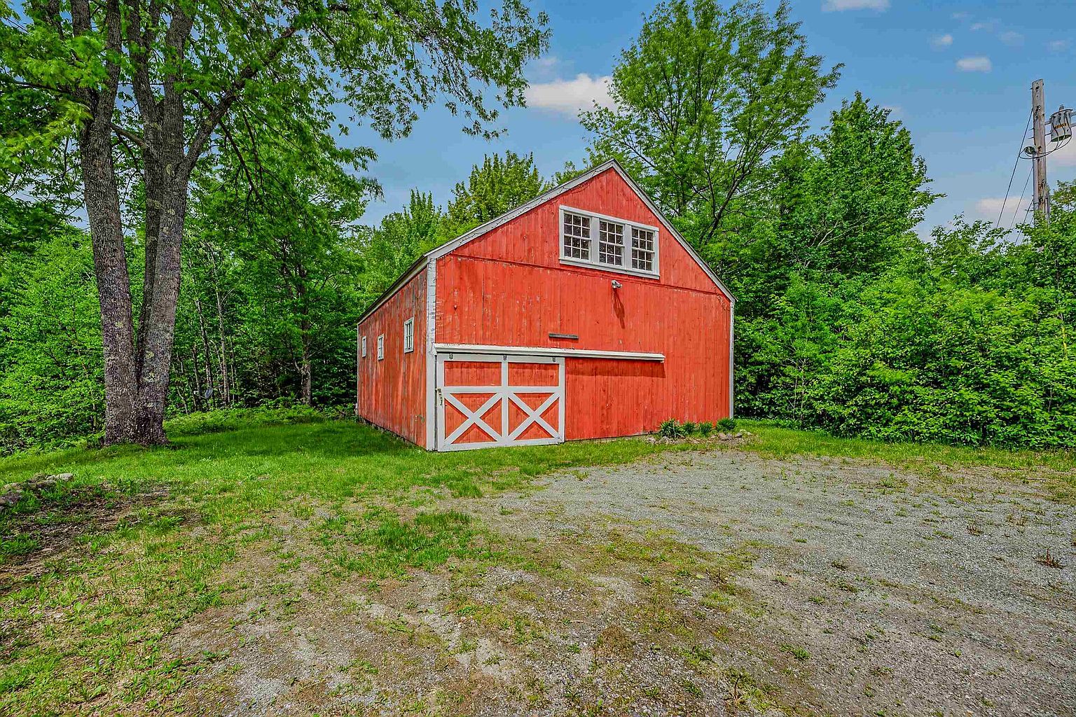 This image captures a classic red barn set against a backdrop of lush green trees under a bright blue sky. The structure features a prominent sliding barn door with white cross-bracing and a large, multi-pane window centered on the upper level. A gravel driveway leads up to the barn, which is surrounded by a well-maintained grassy lawn, creating a rustic and peaceful rural aesthetic.
