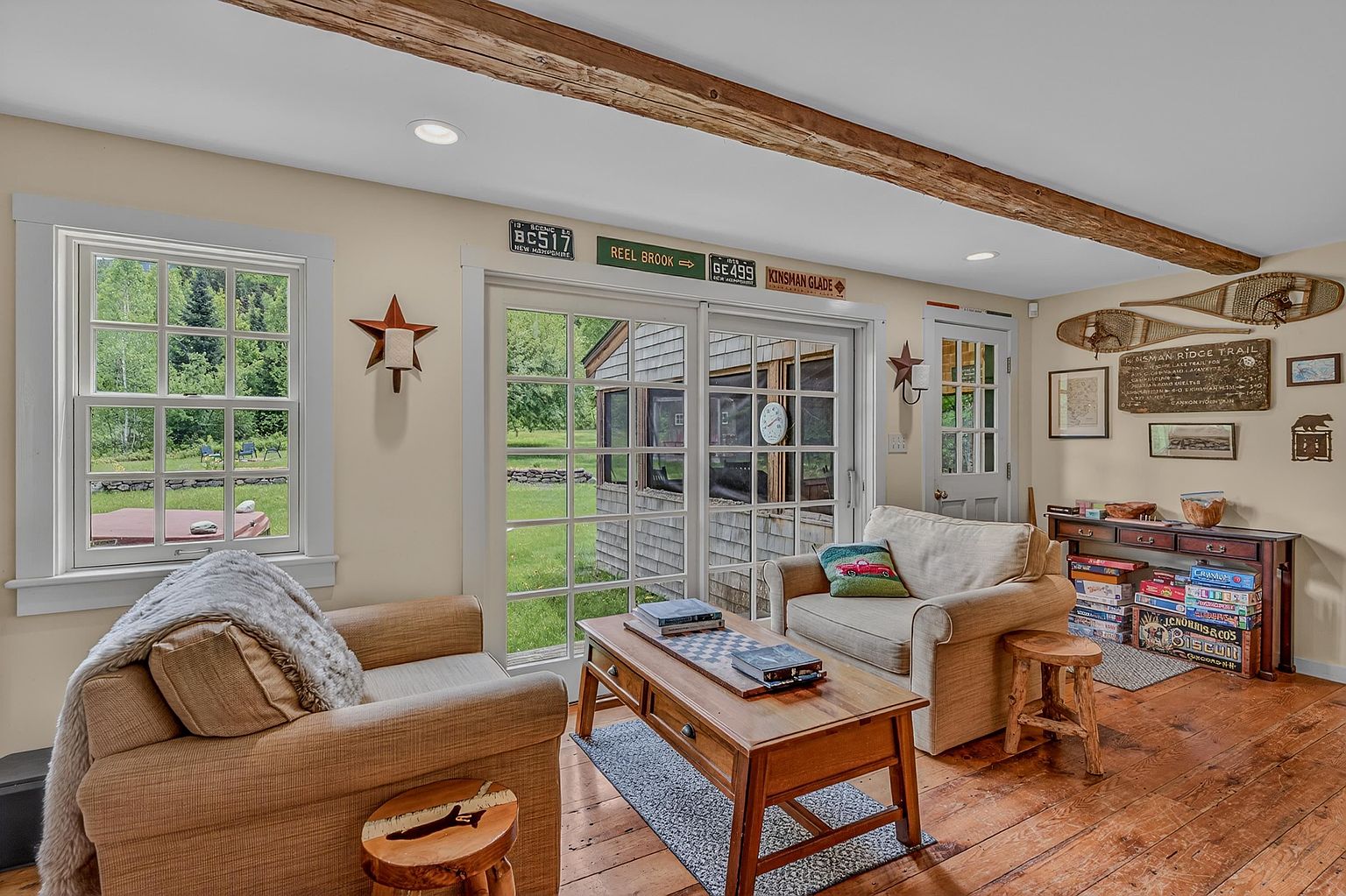 This cozy living room features warm wood flooring, exposed ceiling beams, and a comfortable seating arrangement centered around a wooden coffee table. Large sliding glass doors provide a view of the lush green backyard, while rustic decor elements like snowshoes and vintage-style signs add a charming, cabin-like aesthetic. The space is well-lit by recessed lighting and natural light, creating an inviting and relaxed atmosphere.