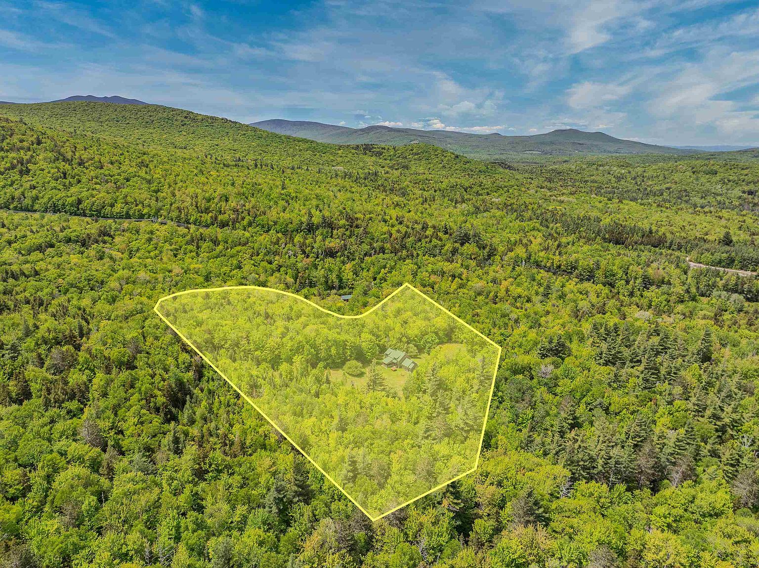This high-angle aerial view captures a secluded property nestled within a dense, lush forest. A yellow polygon overlay highlights the boundaries of the lot, which contains a small house surrounded by mature trees and greenery. The expansive landscape features rolling hills in the background under a bright, partly cloudy sky, emphasizing the property's private and tranquil setting.