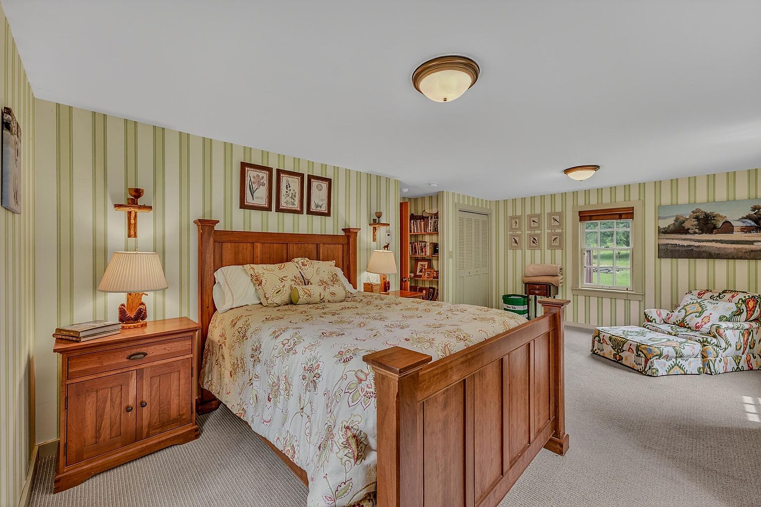 This cozy bedroom features a traditional wooden bed frame as the focal point, complemented by matching nightstands and a patterned floral bedspread. The walls are adorned with vertical striped wallpaper, and a comfortable armchair with an ottoman sits near a window that provides natural light. The room is finished with neutral carpeting and includes built-in shelving and a closet, creating a warm and inviting atmosphere.