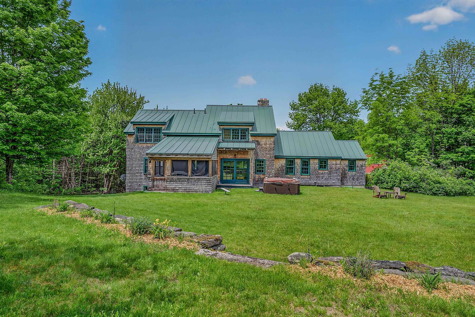 This rear view of a shingle-sided home showcases a classic architectural style with a prominent green metal roof and multiple gables. The property features a screened-in porch, a hot tub situated on the lawn, and a stone retaining wall that separates the manicured grass from the natural landscape. The scene is captured from a wide, eye-level perspective, emphasizing the expansive backyard and the lush, mature trees surrounding the residence.