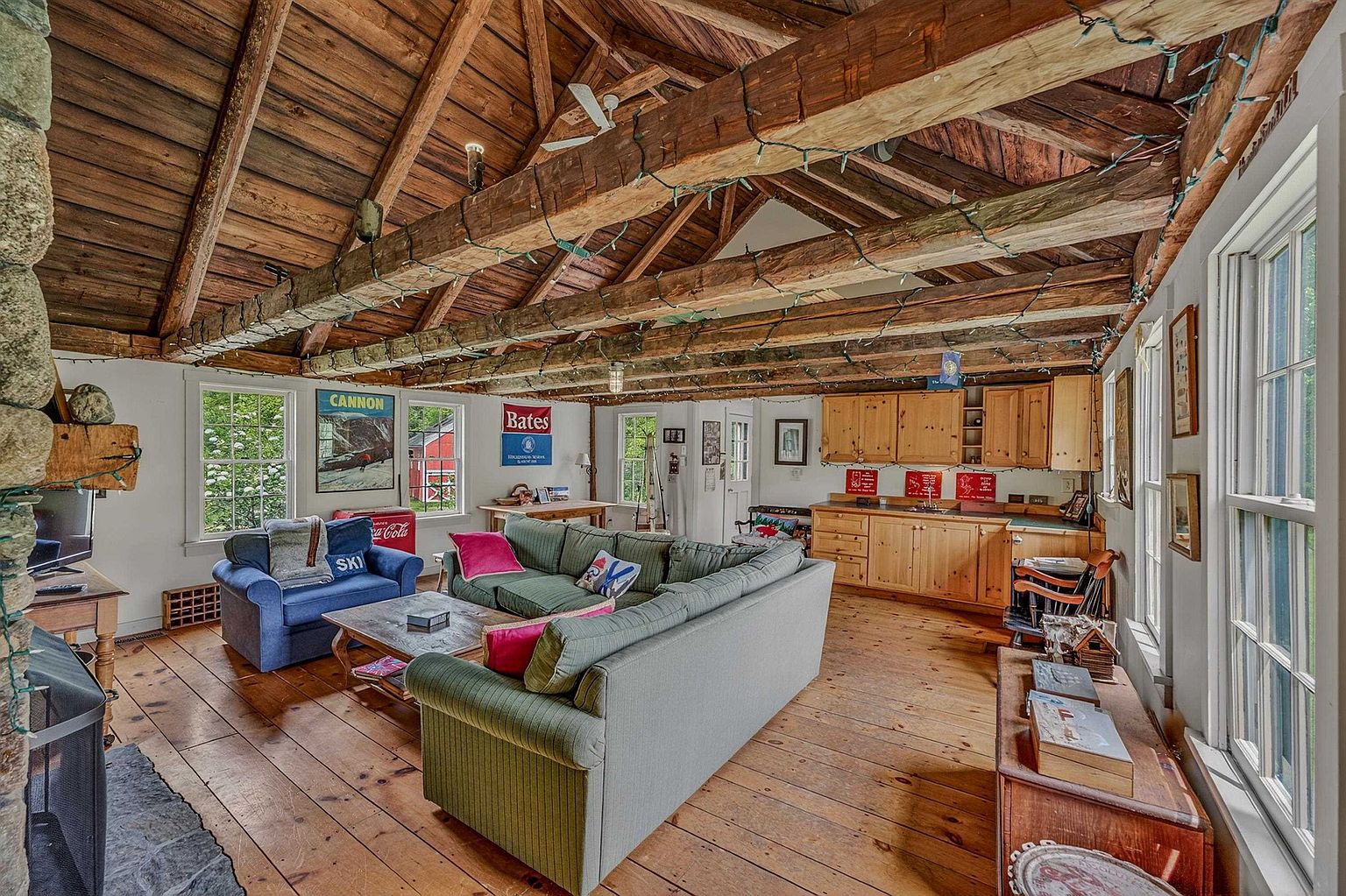 This rustic living room features exposed wooden ceiling beams, wide-plank wood flooring, and a stone-clad chimney breast. The space is furnished with a large green sectional sofa and a blue armchair, creating a cozy, cabin-like atmosphere. A small kitchen area with light wood cabinetry is integrated into the room, while multiple windows provide natural light and views of the surrounding greenery.