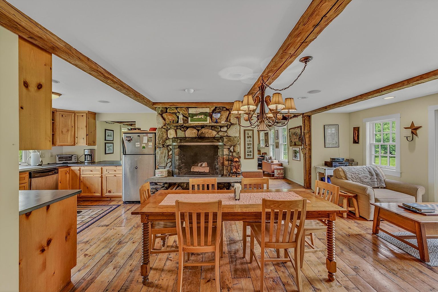 This open-concept living space features a rustic dining area centered around a large wooden table, illuminated by a multi-armed chandelier. A prominent stone-clad chimney breast serves as a focal point between the dining area and the kitchen, which is equipped with light wood cabinetry and stainless steel appliances. The room is characterized by warm wood flooring and exposed ceiling beams, creating a cozy, traditional atmosphere that flows into a comfortable seating area on the right.