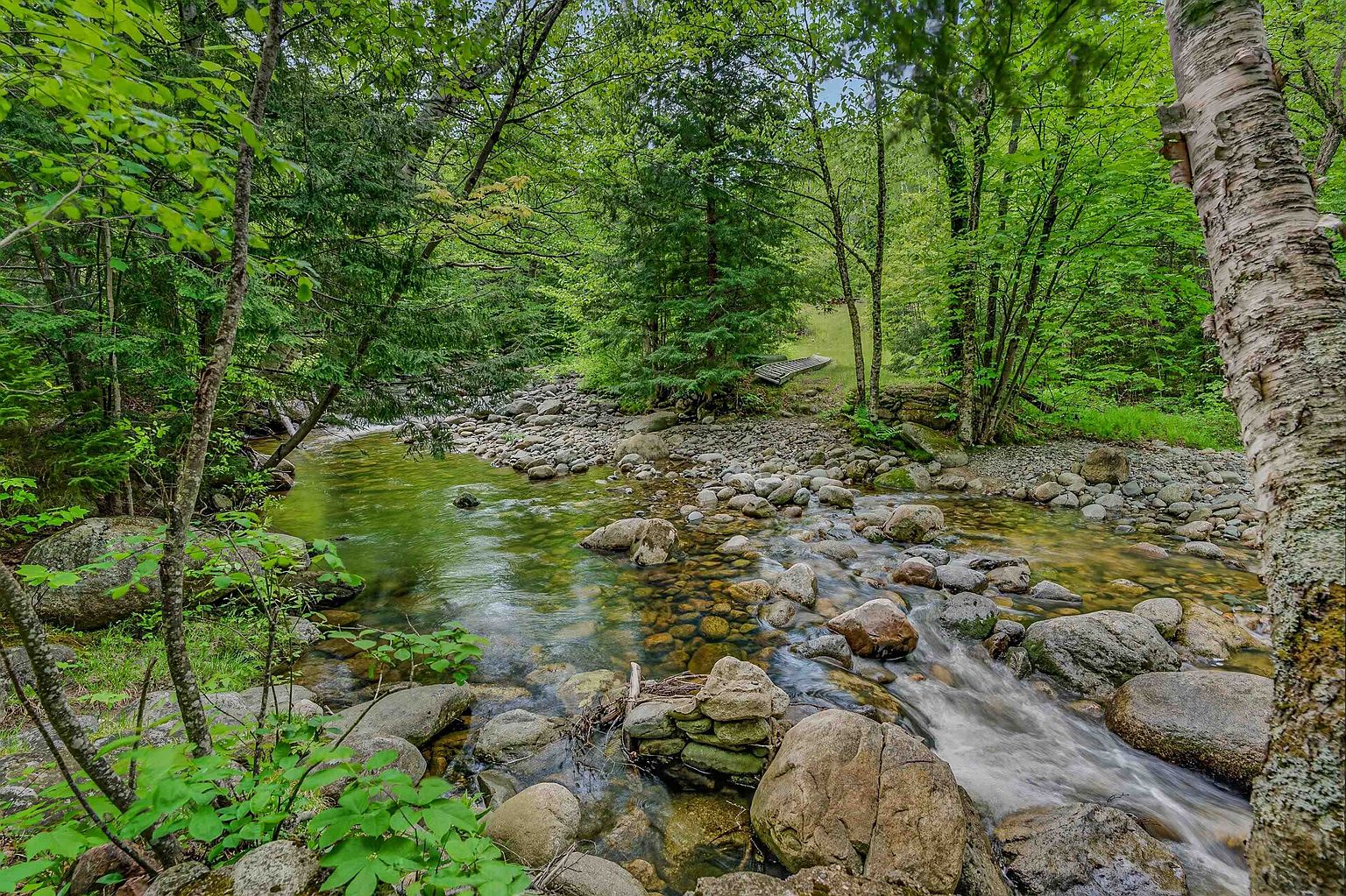 This scenic outdoor space features a tranquil, shallow stream flowing over a bed of smooth river stones, surrounded by a lush, dense forest of mature trees. A small, rustic wooden footbridge is visible in the distance, nestled among the greenery, suggesting a peaceful and private natural setting. The perspective is from the bank of the stream, capturing the serene movement of the water and the vibrant, verdant landscape.