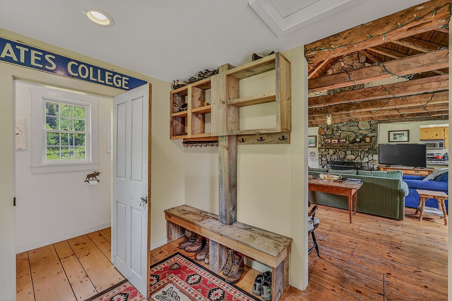 This rustic entryway features a custom-built wooden mudroom bench with integrated shelving and coat hooks, set against light-toned walls and warm wood flooring. A patterned area rug anchors the space, while an open doorway provides a clear line of sight into a cozy living area with exposed timber ceiling beams and a stone-clad chimney breast. The perspective captures a transition between a functional mudroom and a comfortable, cabin-style living space, emphasizing the home's warm and inviting atmosphere.