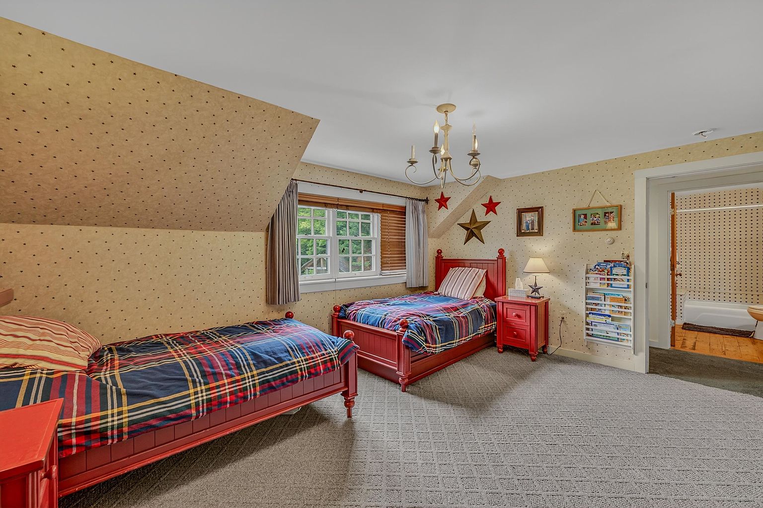 This cozy bedroom features two twin beds with red frames and plaid bedding, set against walls covered in a star-patterned wallpaper. The room includes a central chandelier, a bedside table with a lamp, and a wall-mounted bookshelf, all under a sloped ceiling that adds architectural character. A doorway leads to an adjacent bathroom, and the space is finished with neutral carpeting, creating a warm and inviting atmosphere.