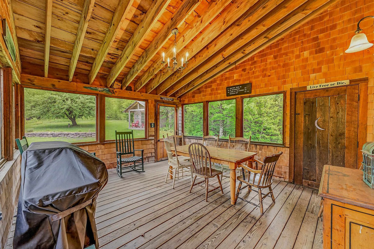 This rustic, screened-in porch features warm wood-paneled walls and a vaulted ceiling with exposed rafters and a classic chandelier. The space is furnished with a wooden dining set and a rocking chair, offering a comfortable area for outdoor-style dining while protected from the elements. Large windows provide expansive views of the surrounding lush green landscape, creating a serene and inviting atmosphere.
