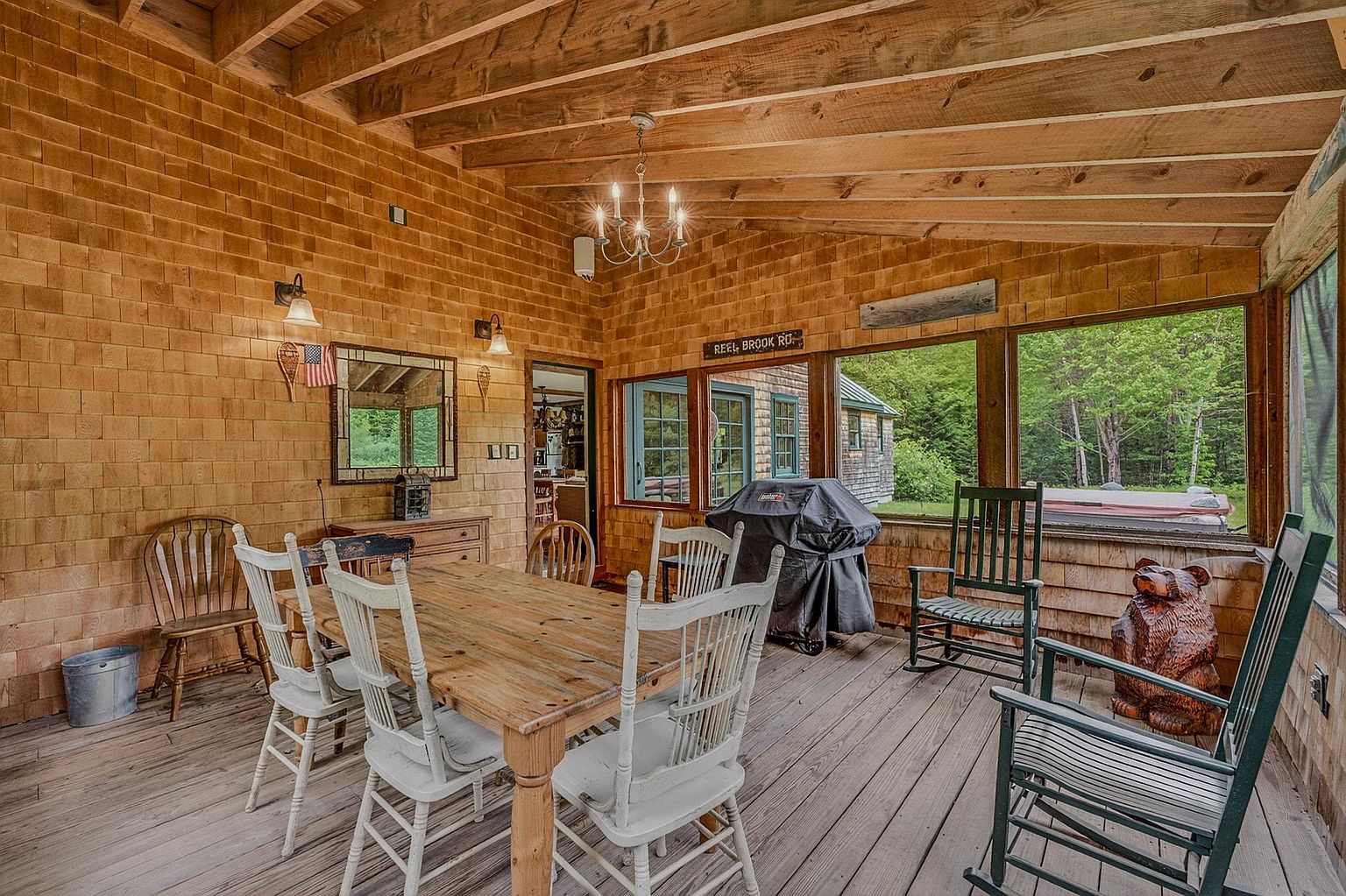 This rustic, screened-in porch serves as a charming dining area, featuring wood-shingled walls and an exposed beam ceiling. The space is furnished with a wooden dining table surrounded by white chairs, complemented by a pair of rocking chairs and a decorative bear carving. Large windows provide a scenic view of the surrounding woods, creating a peaceful and inviting atmosphere for outdoor-style dining.