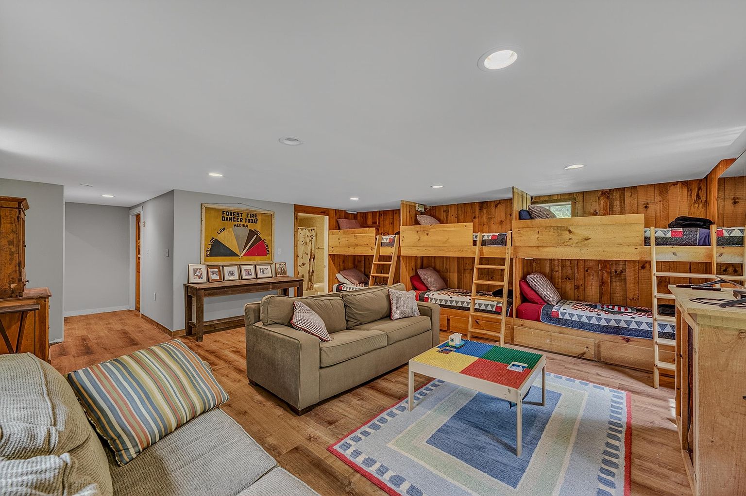 This spacious, rustic-style basement features a cozy living area with a sofa and a colorful coffee table, complemented by a built-in bunk bed unit along the wood-paneled wall. The room is illuminated by recessed lighting and includes a decorative forest fire danger sign, creating a playful, lodge-like atmosphere. The open layout provides ample space for relaxation and sleeping, making it an ideal area for guests or family recreation.
