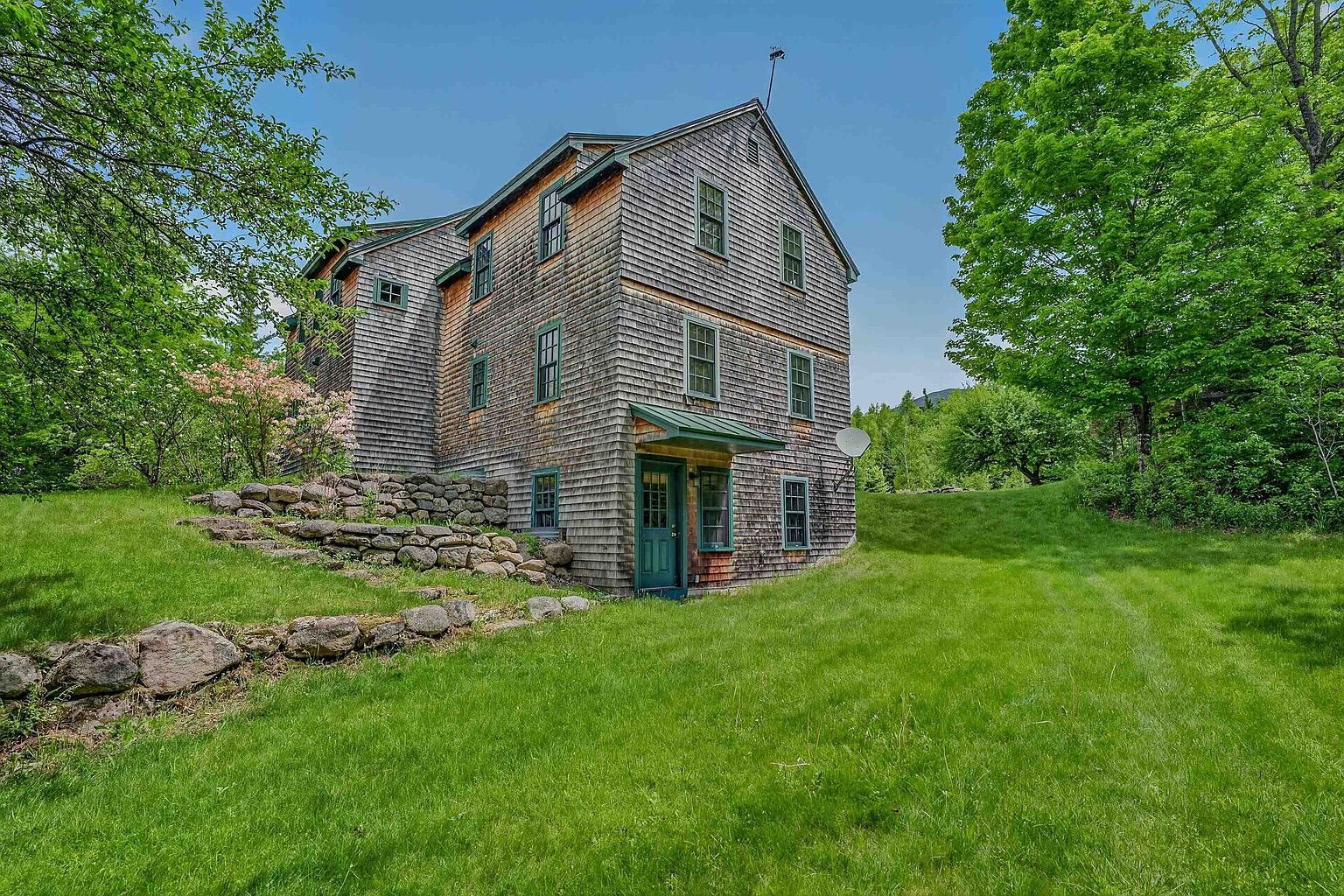 This side view of a multi-story home showcases classic cedar shingle siding and a green-trimmed entryway sheltered by a small metal awning. The property features a tiered stone retaining wall built into the grassy slope, leading up to a lush, tree-lined backyard. The perspective captures the home's rustic charm and its integration into the natural, verdant landscape.