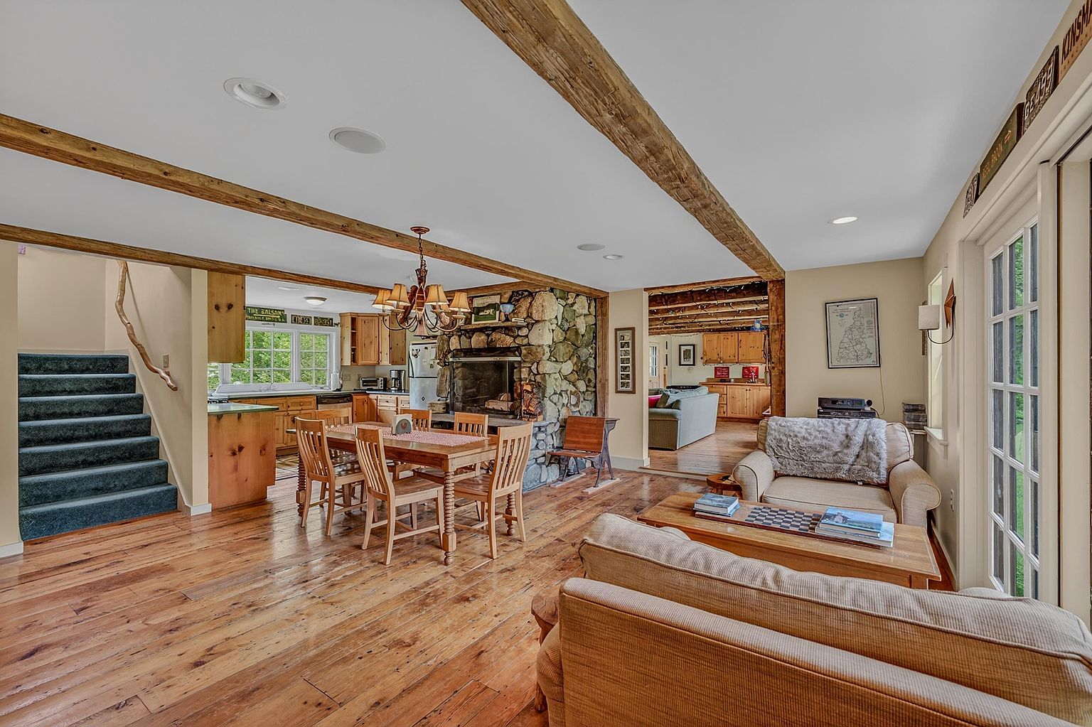 This open-concept living and dining area features warm wood flooring and exposed timber ceiling beams, creating a rustic and inviting atmosphere. A prominent stone-clad chimney breast serves as a central focal point between the dining space and the adjacent kitchen, while a comfortable seating area occupies the foreground. The perspective captures the flow of the home, showcasing a staircase to the left and a view into a secondary living space through a wide doorway.