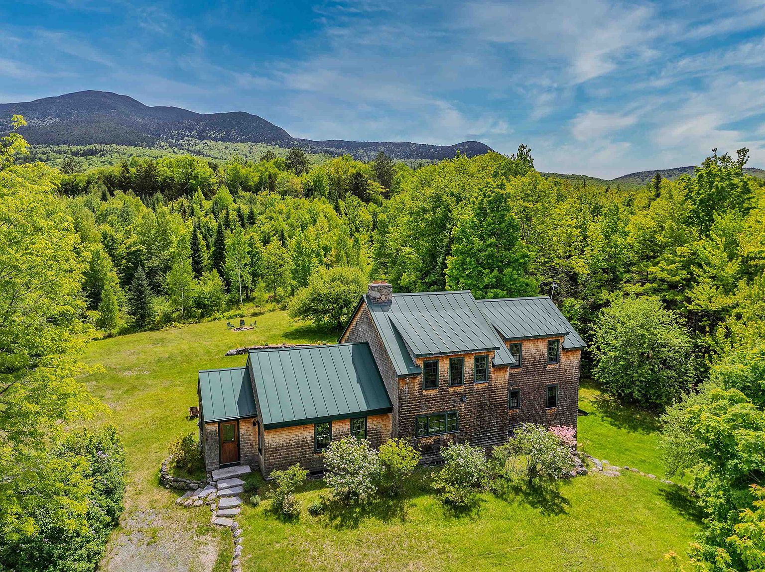 This aerial perspective captures a charming, shingle-sided home nestled within a lush, green landscape against a backdrop of rolling mountains. The house features a distinctive green metal roof, multiple gables, and a stone chimney, blending seamlessly into the surrounding forest. A stone pathway leads to the front entrance, while the expansive lawn and mature trees create a serene and private atmosphere.