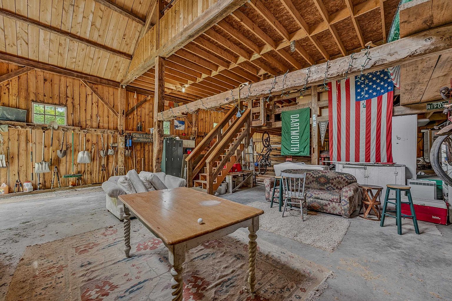 This rustic, barn-style interior space features exposed wooden beams, vertical wood-plank walls, and a concrete floor, creating a casual, multi-purpose recreational area. The room is furnished with a wooden table, two sofas, and a wooden staircase leading to an upper loft level, while tools and sports memorabilia decorate the walls. The wide-angle perspective captures the expansive, open-concept layout, emphasizing the rugged, industrial-farmhouse aesthetic.