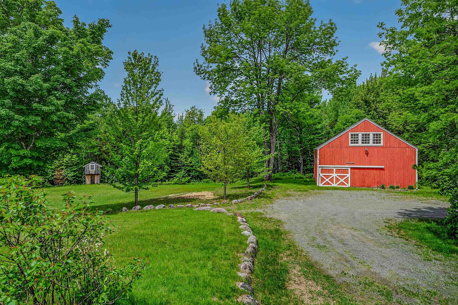 This scenic exterior view features a vibrant red barn with white trim set against a backdrop of lush, mature green trees. A gravel driveway leads toward the barn, while a curved stone border defines a well-maintained grassy lawn in the foreground. A small, rustic wooden outbuilding is nestled among the trees to the left, contributing to the peaceful, rural atmosphere of the property.