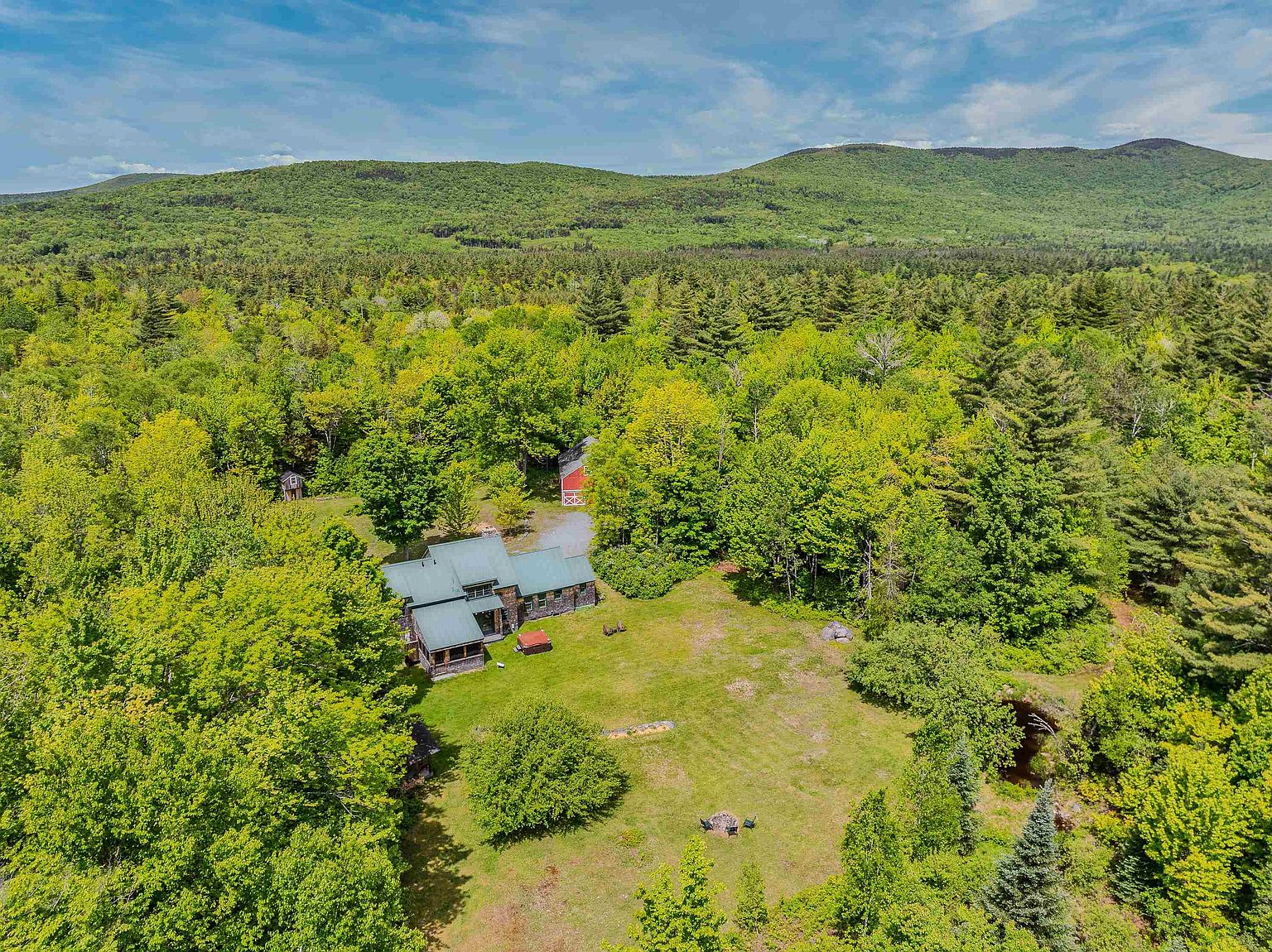 This high-angle aerial perspective captures a secluded property nestled within a lush, forested landscape against a backdrop of rolling mountains. The scene features a multi-sectioned home with a dark roof, a separate red barn, and a spacious grassy yard that includes a fire pit area and a small pond. The expansive greenery and natural setting create a serene, private atmosphere ideal for a rural retreat.