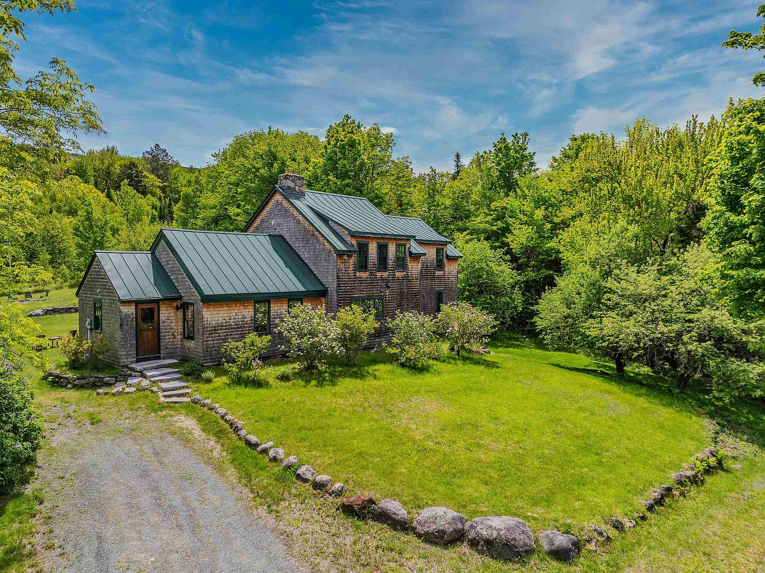 This charming shingle-style home features a distinctive green metal roof and is nestled within a lush, wooded landscape. A gravel driveway leads to the front entrance, which is accented by stone steps and surrounded by mature greenery and a manicured lawn. The home's rustic aesthetic is complemented by its natural setting, creating a peaceful and inviting atmosphere.