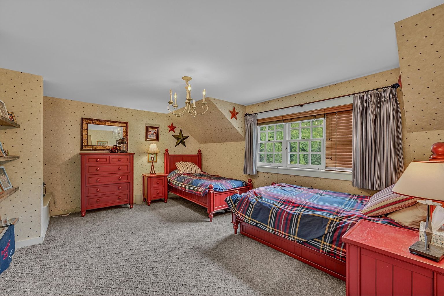 This charming bedroom features two twin beds with plaid bedding, set against walls adorned with a classic star-patterned wallpaper. The room includes a red wooden dresser, matching nightstands, and a central chandelier, creating a cozy, traditional aesthetic. A large window with wooden blinds allows natural light to fill the space, while the sloped ceiling adds architectural character.