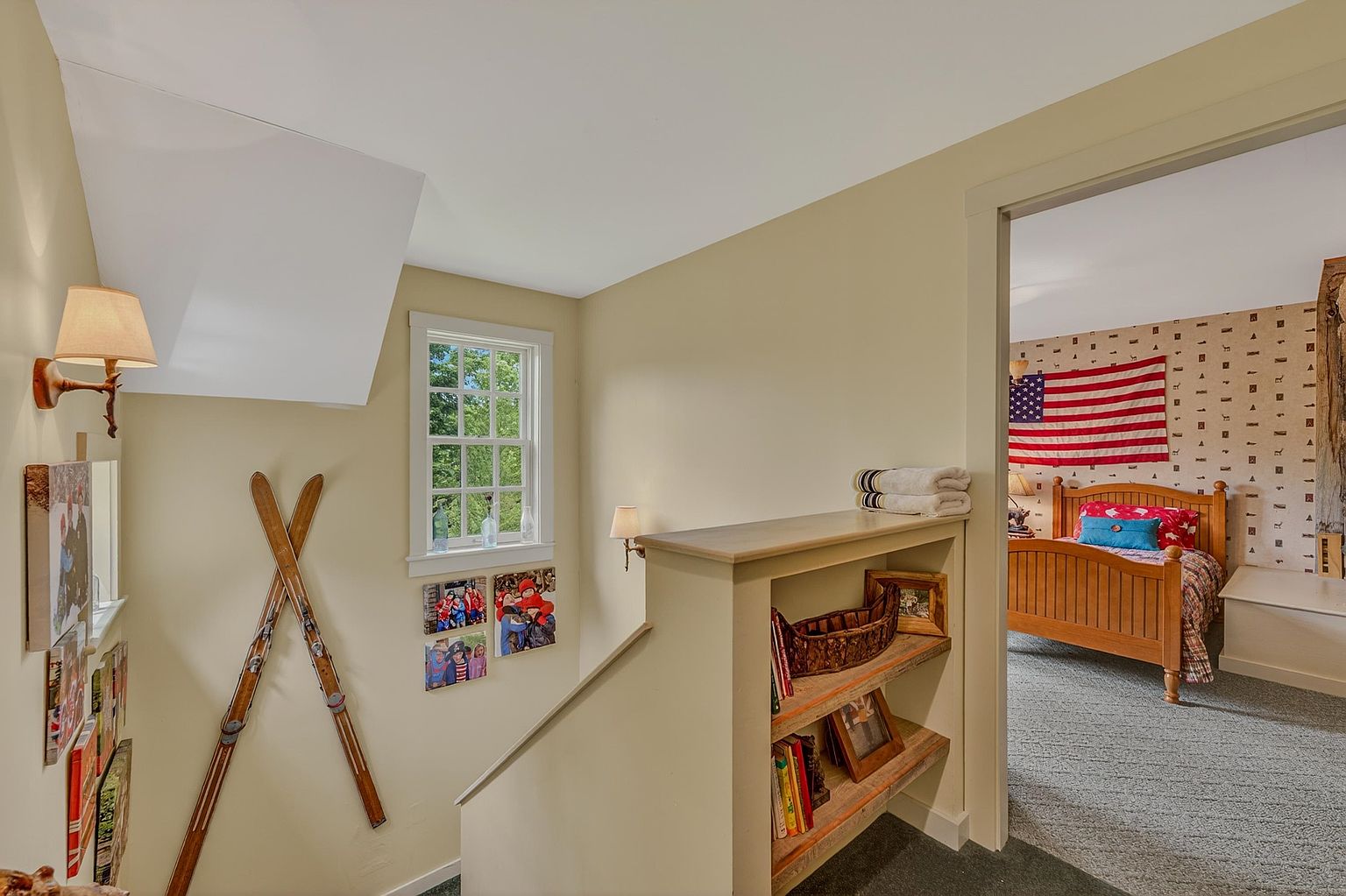This interior view captures a cozy landing area featuring a built-in wooden bookshelf and a staircase adorned with decorative vintage skis. The neutral-toned walls are decorated with framed photos, leading the eye toward an open doorway that reveals a bedroom with an American flag wall hanging. The perspective provides a warm, inviting transition between levels, highlighting the home's rustic and personalized character.