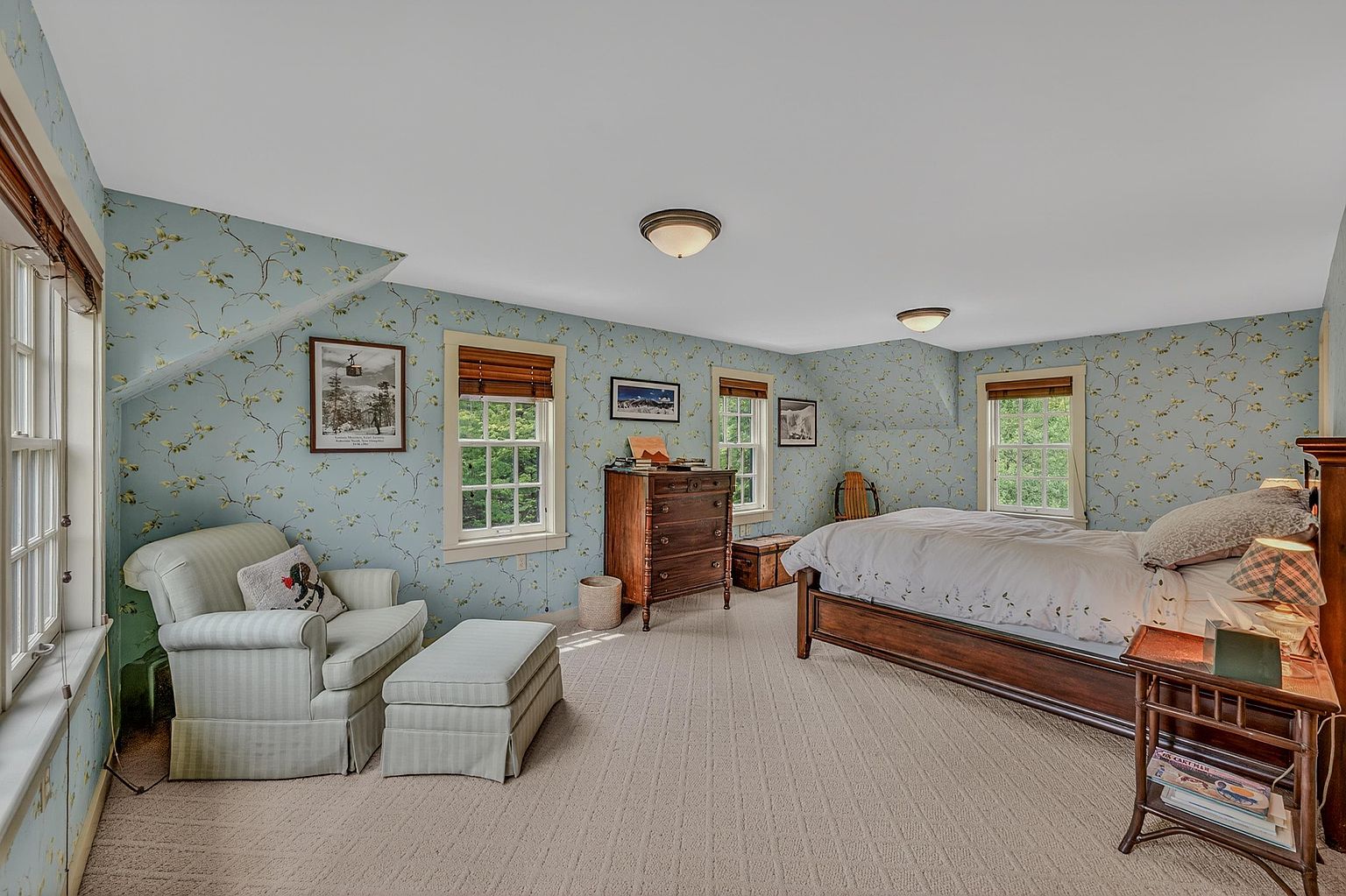 This cozy bedroom features charming floral wallpaper and a neutral carpet, creating a warm and inviting atmosphere. The room is furnished with a wooden bed frame, a matching dresser, and a comfortable armchair with an ottoman placed near the windows. The space is well-lit by natural light streaming through multiple windows, highlighting the traditional decor and sloped ceiling architecture.