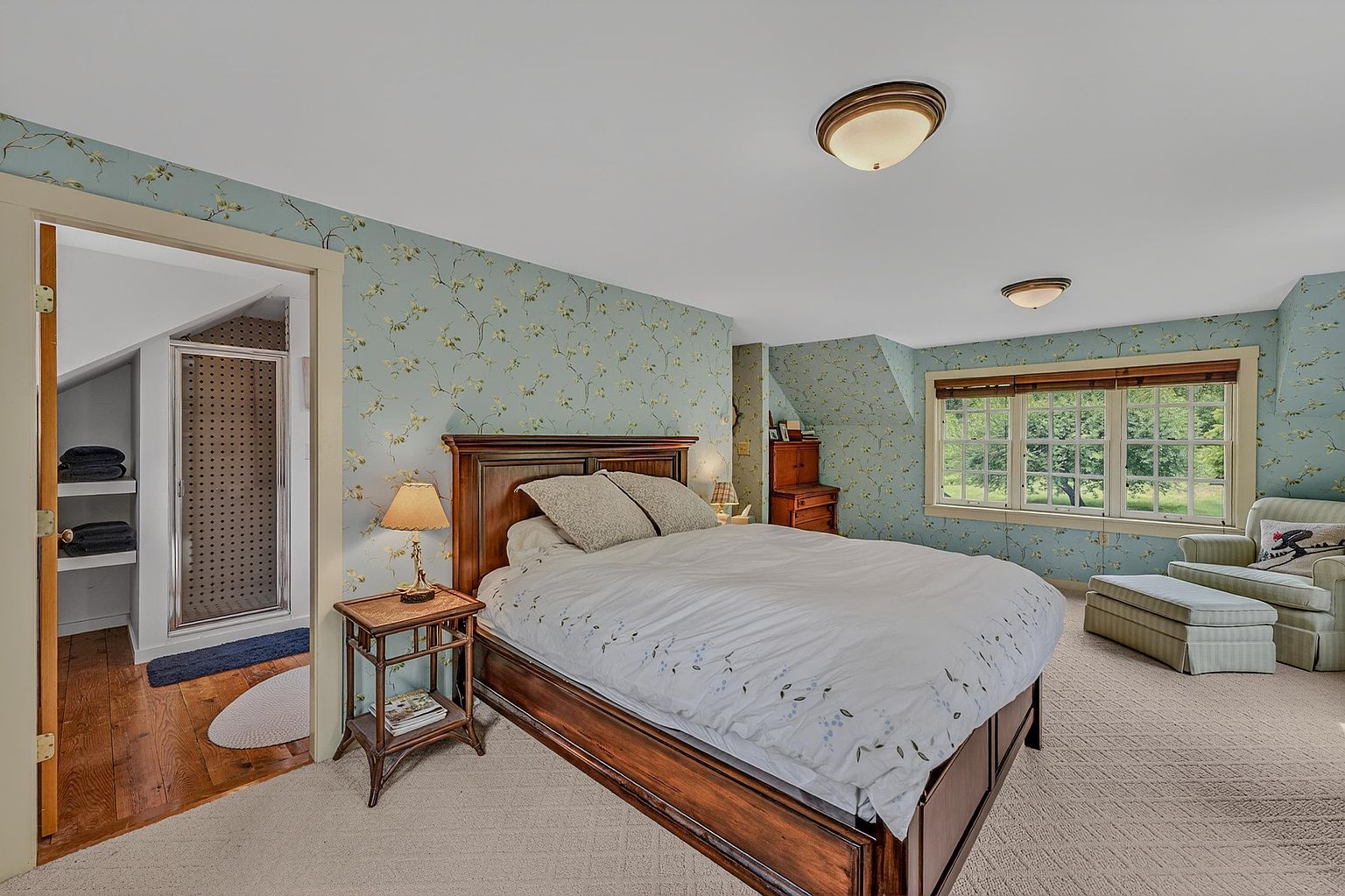 This primary bedroom features a classic aesthetic with light blue floral wallpaper and a large wooden bed frame as the central focal point. A cozy sitting area with an armchair and ottoman is positioned near a set of windows that provide natural light and views of the outdoors. To the left, an open doorway reveals a glimpse into an adjoining bathroom with a glass-enclosed shower and wooden flooring.