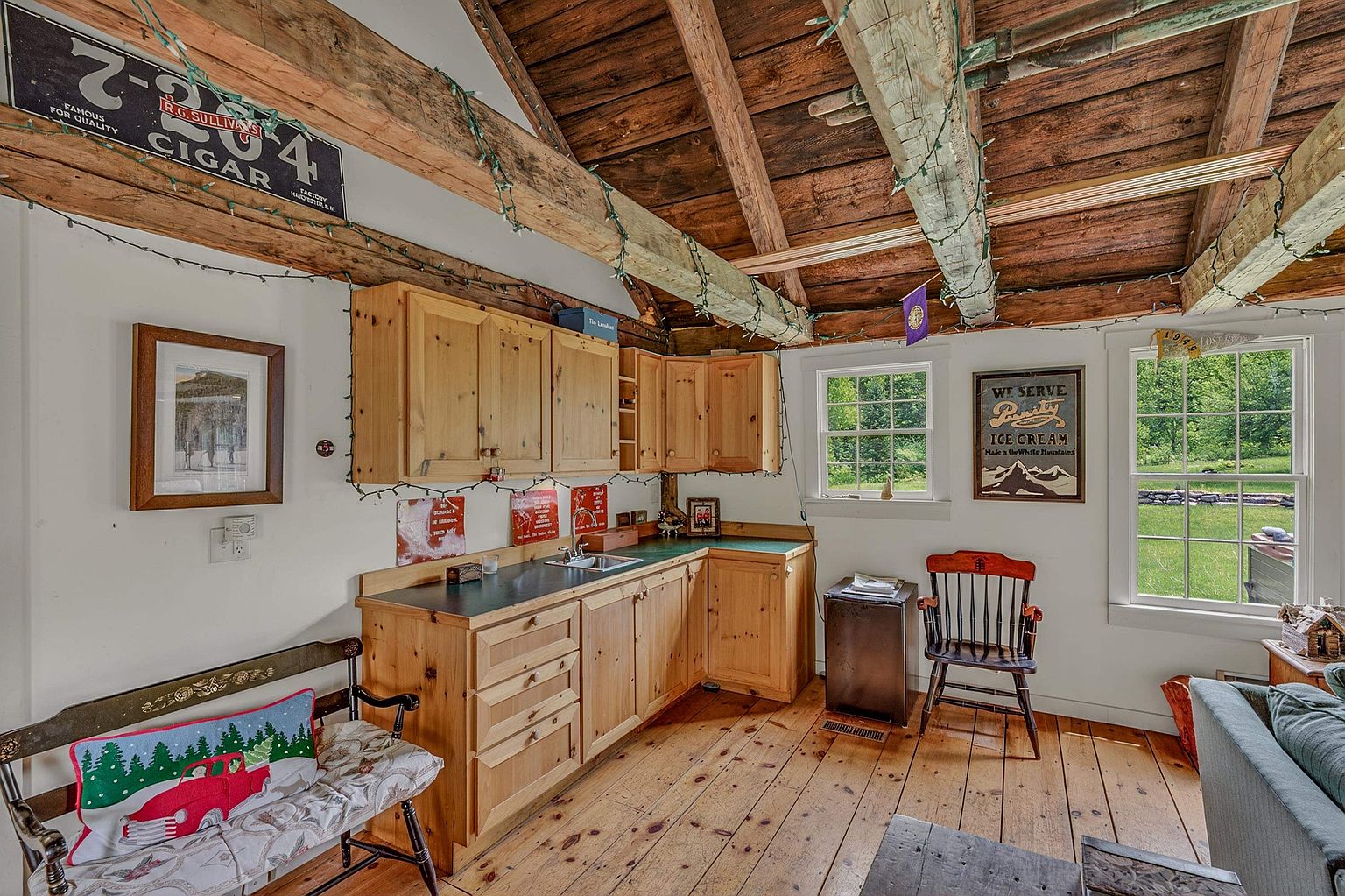This rustic kitchen features light wood cabinetry, a dark countertop, and exposed wooden ceiling beams that add character to the space. The room is decorated with vintage-style signage and a wooden bench, creating a cozy, cabin-like atmosphere. Large windows allow natural light to fill the room, highlighting the warm wood flooring and the charming, eclectic decor.