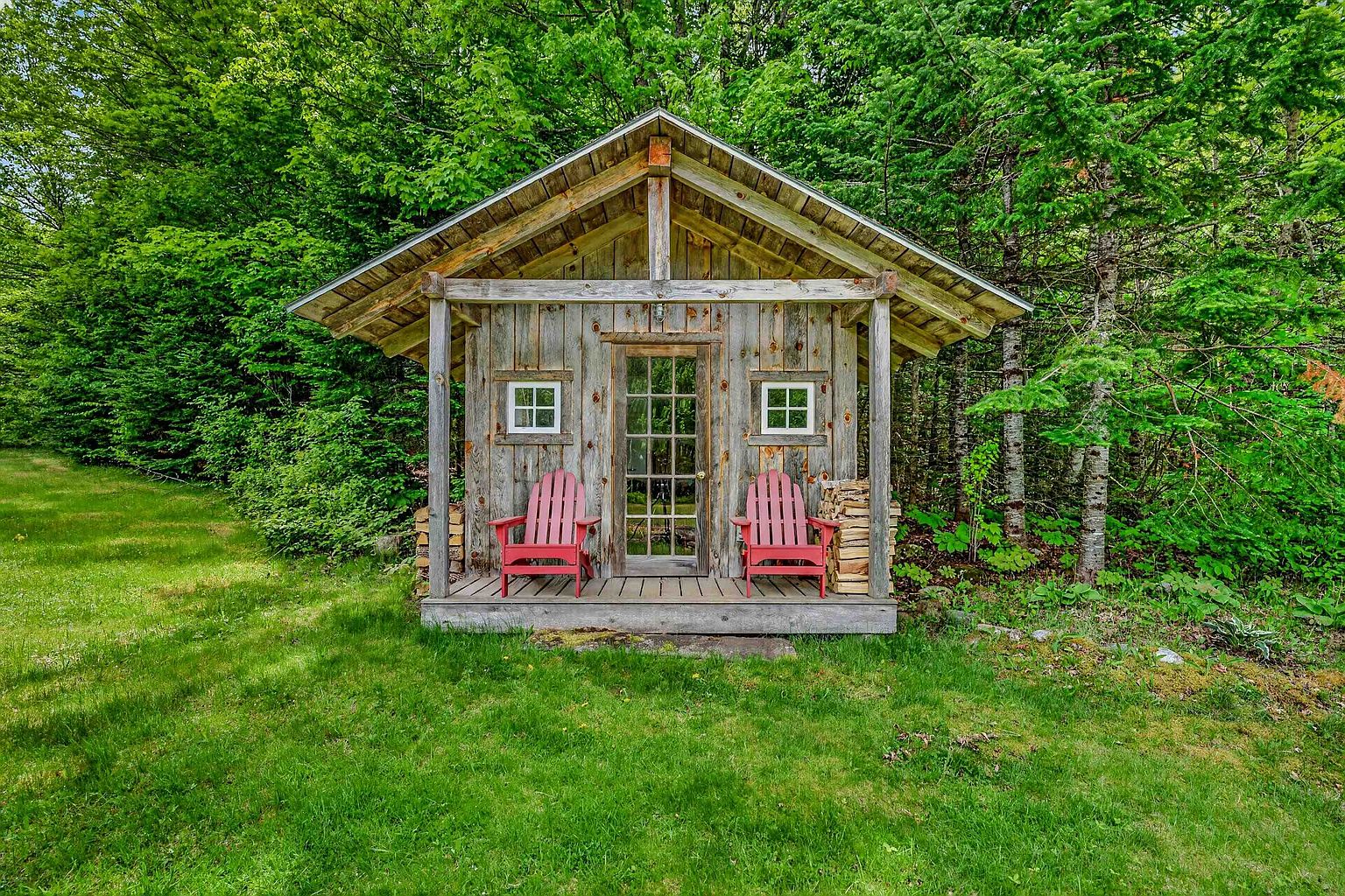 This charming, rustic wooden cabin features a covered front porch with two red Adirondack chairs, creating a cozy and inviting outdoor seating area. The structure is built with vertical wood siding and is framed by a lush, dense forest backdrop, emphasizing a peaceful, secluded setting. A central glass-paned door and two small windows provide symmetry to the facade, while stacks of firewood are neatly stored on either side of the porch.