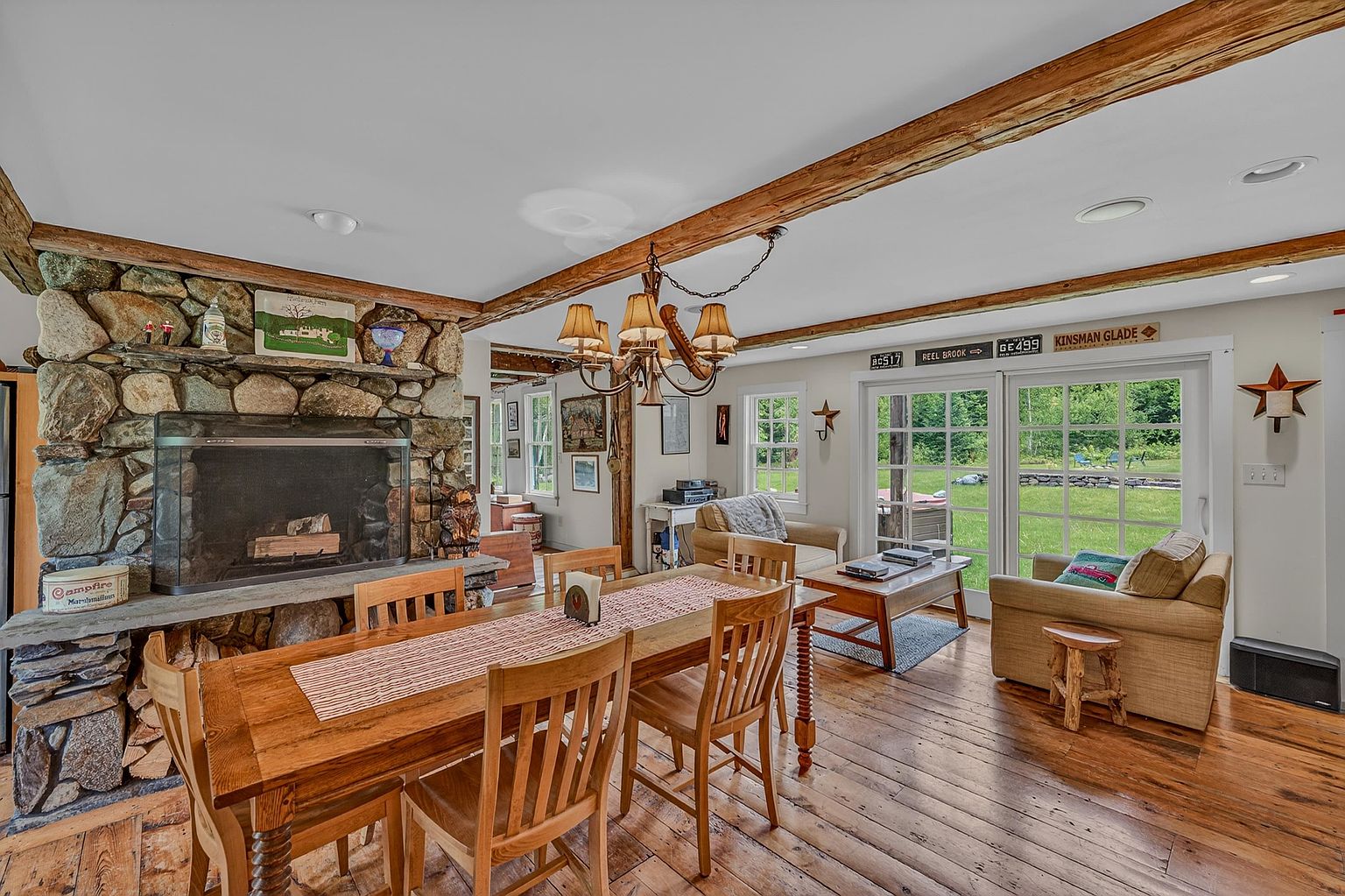 This rustic dining area features a large wooden table as its centerpiece, positioned in front of a prominent stone-clad chimney breast with an integrated fireplace. The room showcases exposed wooden ceiling beams and warm-toned wood flooring, creating a cozy, cabin-like atmosphere. Large glass doors provide a clear view of the lush green backyard, while a traditional chandelier hangs above the dining table to complete the inviting space.