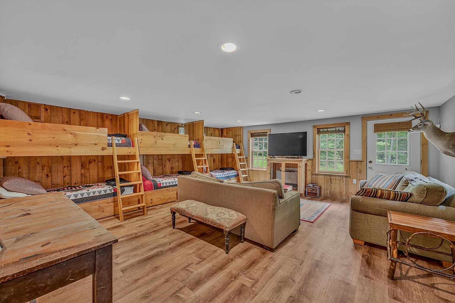 This spacious, rustic-style bunk room features multiple built-in wooden bunk beds along one wall, providing ample sleeping accommodations. The room includes a comfortable seating area with a sofa and a bench, a television, and wood-look flooring, creating a cozy and functional retreat. The perspective is wide-angle, capturing the entire layout of the room, including the wood-paneled walls and the taxidermy mount near the entryway.