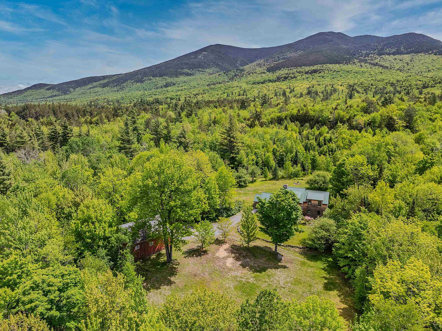 This high-angle aerial view captures a secluded property nestled within a lush, dense forest at the base of a prominent mountain range. The scene features a rustic home and a separate red barn, both surrounded by expansive green lawns and mature trees. The perspective emphasizes the property's expansive natural setting and tranquil, private atmosphere.