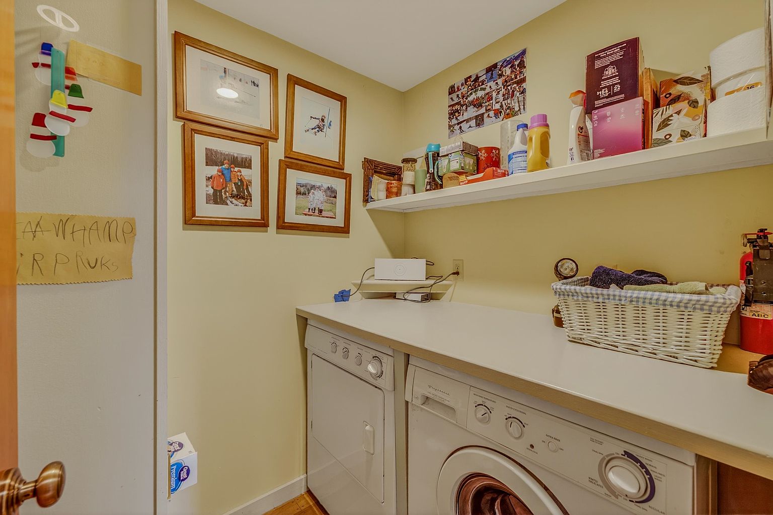 This functional laundry room features a side-by-side washer and dryer set topped with a white counter for folding. Above the appliances, a wall-mounted shelf provides storage for cleaning supplies and household items, while framed family photos decorate the adjacent wall. The space is painted in a soft yellow tone, creating a warm and practical utility area.