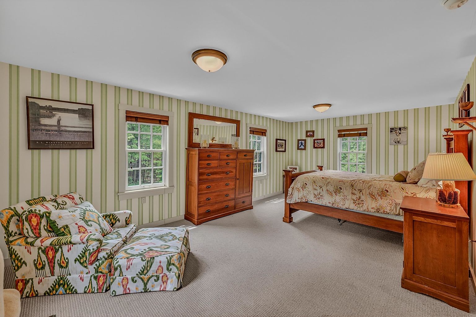 This spacious bedroom features light green and white striped wallpaper and neutral carpeting, creating a traditional and cozy atmosphere. The room is furnished with a wooden bed frame, a matching dresser with an integrated mirror, and a patterned armchair with a coordinating ottoman. Large windows allow natural light to fill the space, highlighting the classic decor and framed wall art.