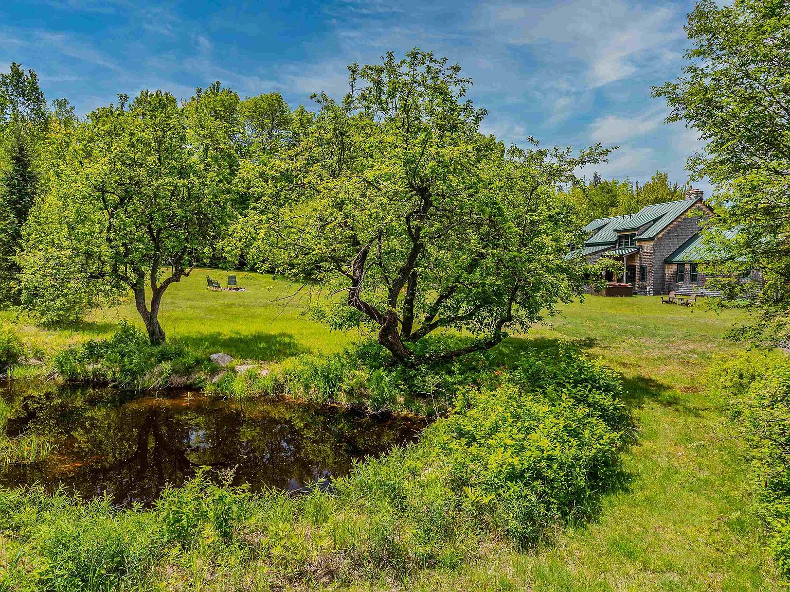 This scenic exterior view captures a lush, expansive green lawn surrounding a tranquil pond in the foreground. A charming shingled house with a green metal roof sits nestled among mature trees, creating a peaceful and private atmosphere. The perspective is a wide-angle shot that emphasizes the natural beauty and generous outdoor space of the property.