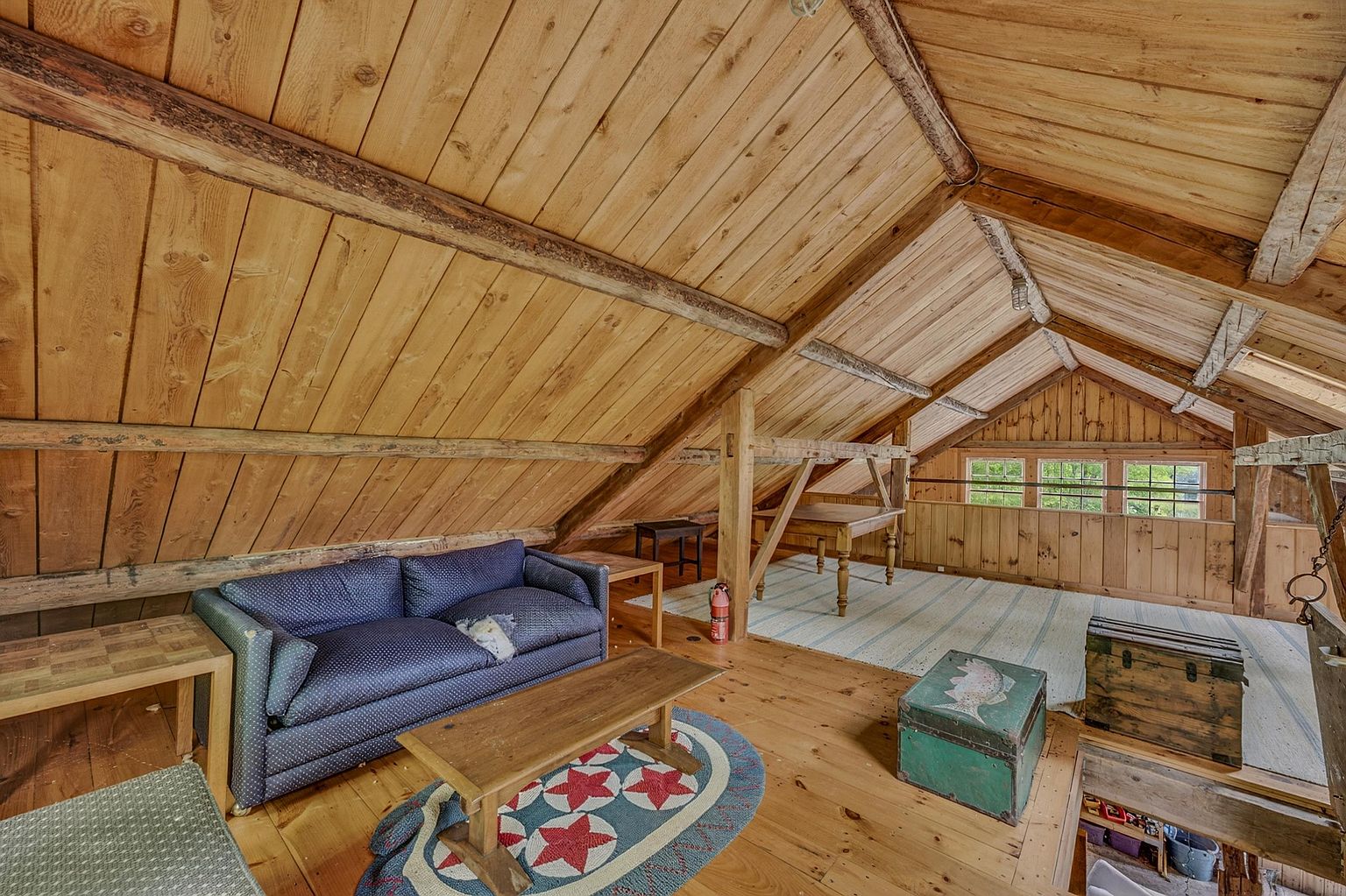 This rustic attic space features exposed wooden beams and plank walls, creating a warm, cabin-like atmosphere. The room is furnished with a blue patterned sofa, a wooden coffee table on a star-motif rug, and a dining table set further back near a row of windows. The open-concept layout under the pitched roof provides a cozy, versatile living area with a distinct vintage charm.