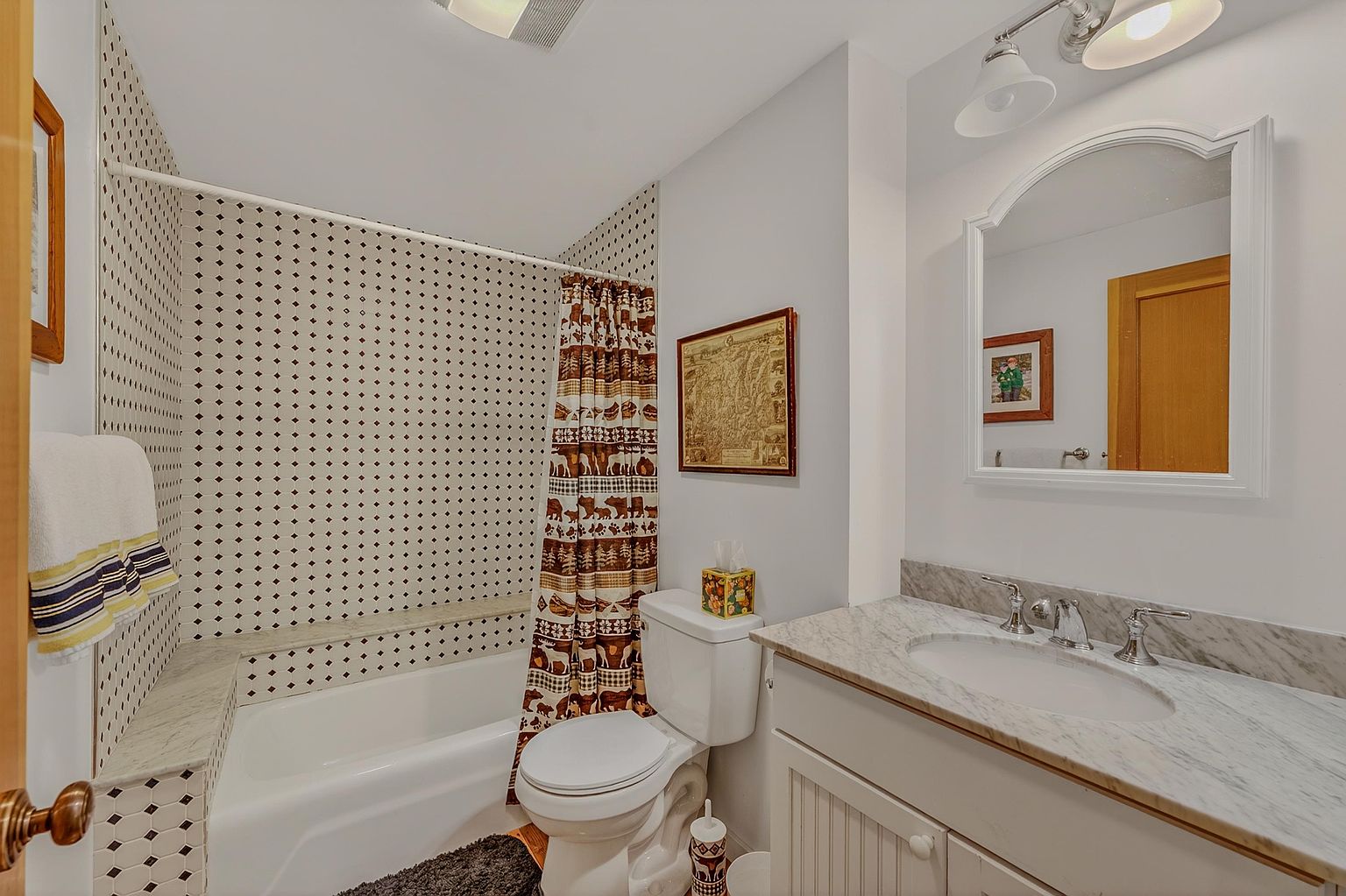 This bathroom features a vanity with a stone countertop and an arched mirror, complemented by a shower-tub combination enclosed by a patterned shower curtain. The walls are adorned with a distinctive diamond-patterned tile surround in the bathing area, while the rest of the room is painted in a neutral tone. A framed map hangs on the wall above the toilet, adding a decorative touch to the compact and functional space.