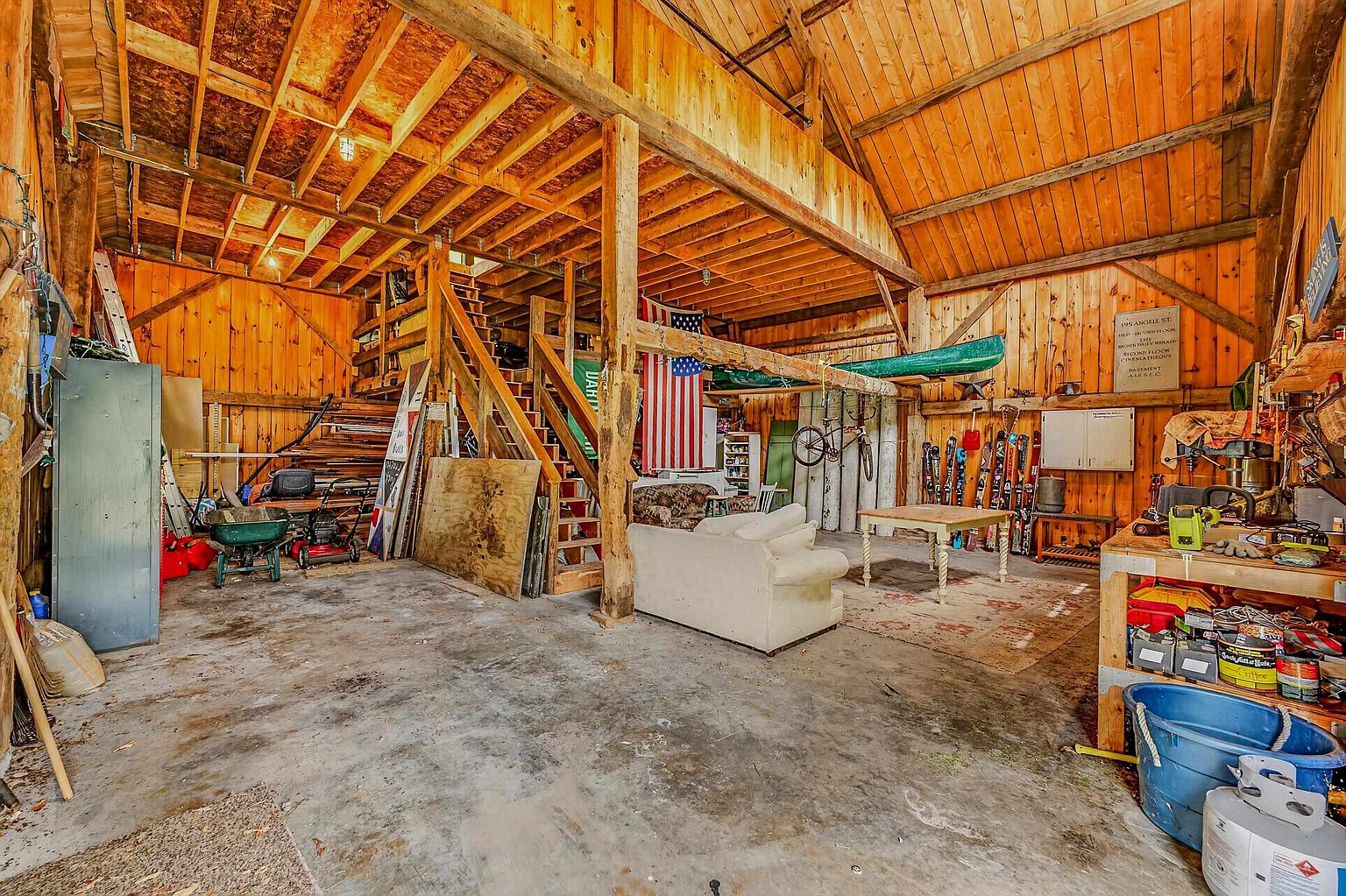 This expansive, rustic barn-style garage features exposed timber framing, high vaulted ceilings, and a concrete floor. The space is multi-functional, serving as both a storage area for tools and equipment and a casual lounge with a sofa and table, while a wooden staircase leads to an upper level. A green canoe is suspended from the rafters, and various recreational items like skis and a bicycle are mounted on the walls, creating a versatile and hobby-focused environment.