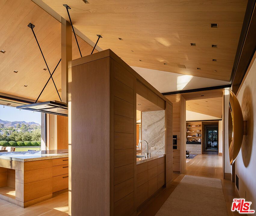 This sophisticated, modern kitchen features a central wooden island partition that separates the cooking area from the hallway, showcasing high-end craftsmanship and clean lines. The space is defined by warm, light-toned wood cabinetry and ceilings, complemented by a marble backsplash and a sleek, industrial-style range hood. The perspective looks through the kitchen toward a bright, open hallway, emphasizing the seamless flow and airy, sun-drenched atmosphere of the home.