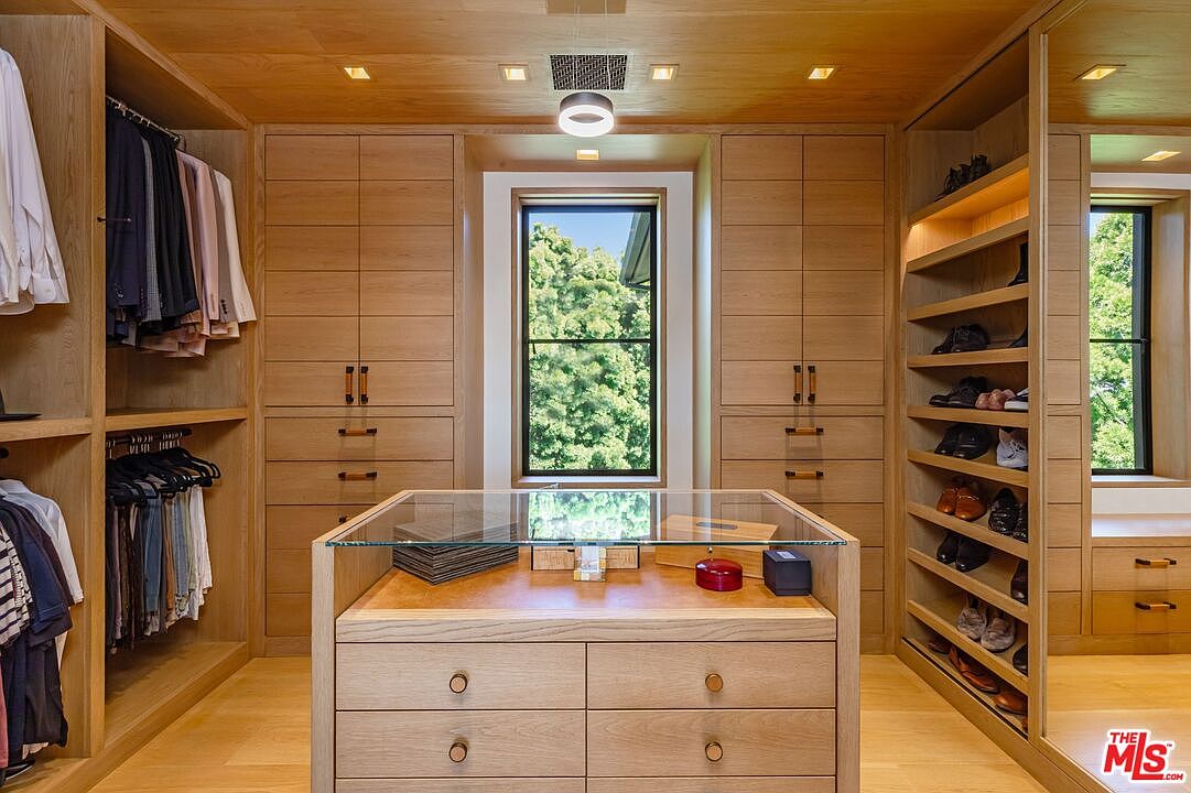 This luxurious, custom-designed walk-in closet features warm, light-toned wood cabinetry throughout, providing extensive storage for clothing and accessories. A central island with a glass top serves as a display case, while a centered window allows natural light to brighten the space. The symmetrical layout and integrated shelving create a sophisticated, organized, and high-end aesthetic.