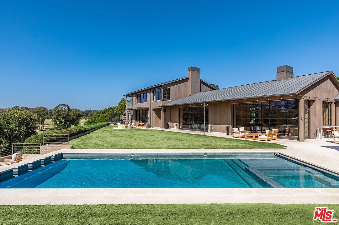 This stunning rear exterior view showcases a modern, custom-designed swimming pool and integrated spa set against a backdrop of a sprawling, manicured lawn and a contemporary wood-sided residence. The home features expansive floor-to-ceiling glass walls that seamlessly blend indoor and outdoor living spaces, complemented by a sleek metal roof. The perspective captures the serene, resort-like atmosphere of the property, highlighting the clean lines and high-end architectural design.
