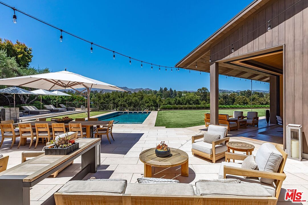 This expansive outdoor living space features a seamless transition from a covered patio to a sun-drenched pool deck, perfect for luxury entertaining. The area is furnished with high-end wooden lounge seating, a large dining table under a modern umbrella, and a pristine swimming pool overlooking a lush green lawn and distant hills. String lights overhead add a warm, inviting ambiance to this sophisticated, resort-style backyard.