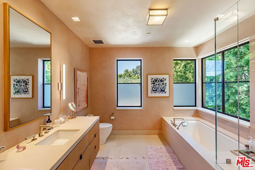 This elegant primary bathroom features a warm, earthy color palette with textured plaster walls and a sleek wooden vanity. The space is anchored by a deep soaking tub positioned beneath large windows that offer a serene view of the surrounding greenery, complemented by a modern glass-enclosed shower area. A large, minimalist mirror and contemporary lighting fixtures enhance the bright, airy, and sophisticated atmosphere of the room.