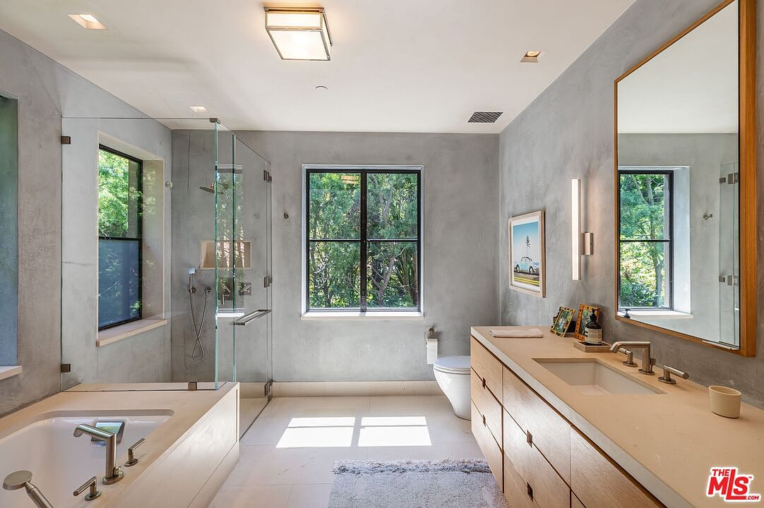 This modern primary bathroom features a sleek, minimalist aesthetic with light gray concrete-style walls and warm wood cabinetry. The space includes a spacious walk-in glass shower, a deep soaking tub, and a large vanity with a wide mirror, all bathed in natural light from the windows overlooking lush greenery. The clean lines and neutral color palette create a serene, spa-like atmosphere.