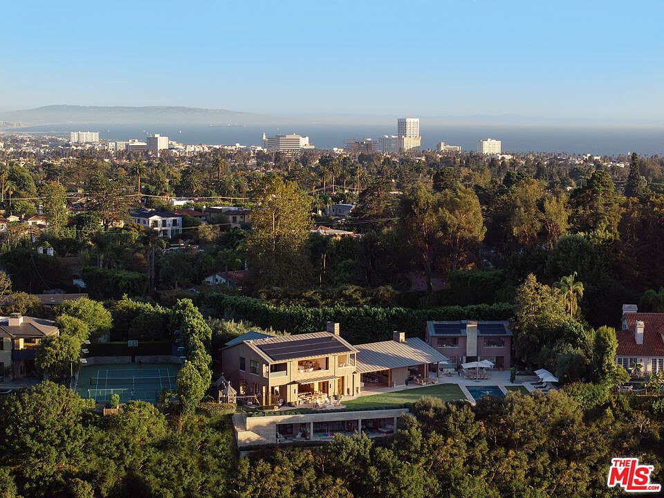 This high-angle aerial shot captures a luxurious residential property nestled within a lush, tree-filled landscape, offering a panoramic view of the distant coastline and city skyline. The home features a modern architectural design with solar panels on the roof, a private tennis court, and a swimming pool area, all integrated seamlessly into the verdant surroundings. The perspective emphasizes the expansive scale of the estate and its prime location, blending privacy with breathtaking vistas.