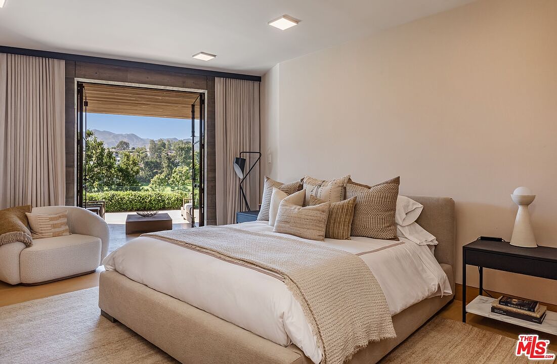 This elegant primary bedroom features a neutral color palette, a plush upholstered bed, and a cozy curved armchair, creating a serene and sophisticated atmosphere. Large glass doors open onto a private patio, seamlessly blending indoor living with the scenic outdoor landscape. The room is accented by modern lighting fixtures and minimalist decor, emphasizing a luxurious and tranquil retreat.
