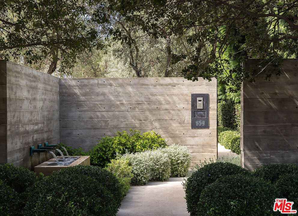 This sophisticated entryway features a striking board-formed concrete wall that provides a modern, textured backdrop for a minimalist water feature. Lush, manicured greenery and rounded shrubs frame a clean concrete pathway, leading visitors toward a discreet, integrated intercom system. The overall aesthetic is one of serene, contemporary luxury, emphasizing privacy and architectural precision.