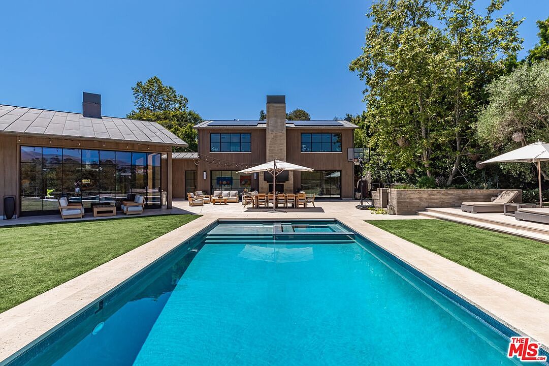 This stunning backyard features a large, rectangular swimming pool as the centerpiece, flanked by manicured green lawns and expansive stone patio areas. The modern, multi-story home serves as a dramatic backdrop, showcasing floor-to-ceiling glass walls, a prominent stone chimney, and an outdoor dining area shaded by a large umbrella. The scene captures a luxurious, resort-like atmosphere perfect for high-end outdoor entertaining.