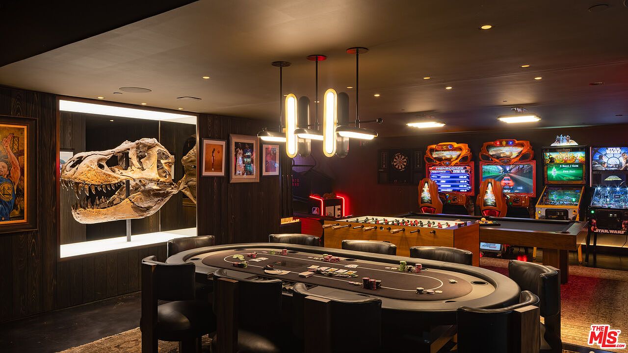 This luxurious entertainment room features a central poker table surrounded by leather chairs, set against a backdrop of a dramatic, illuminated T-Rex skull display. The space is equipped with multiple arcade cabinets, a foosball table, and a dartboard, creating a high-end game room atmosphere. The dark wood paneling and modern lighting fixtures contribute to a sophisticated, masculine aesthetic perfect for hosting.