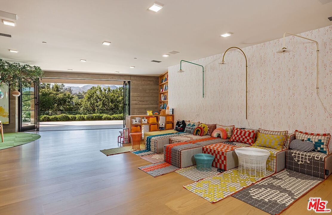 This spacious, modern playroom features light hardwood flooring and a large, open-concept design that seamlessly connects to an outdoor patio through expansive folding glass doors. The room is furnished with colorful, modular floor seating and patterned rugs, creating a vibrant and playful atmosphere. A built-in wooden bookshelf and whimsical wall-mounted lighting fixtures add character to the space, making it an ideal area for family activities.