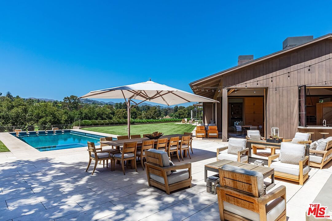 This expansive outdoor patio area features a large dining table under a modern umbrella, surrounded by comfortable wooden lounge seating with plush cushions. The space seamlessly transitions from the home's interior to a pristine swimming pool and a manicured lawn, offering a perfect setting for luxury entertaining. The architectural style is contemporary and warm, emphasizing an indoor-outdoor lifestyle with stunning views of the surrounding landscape.